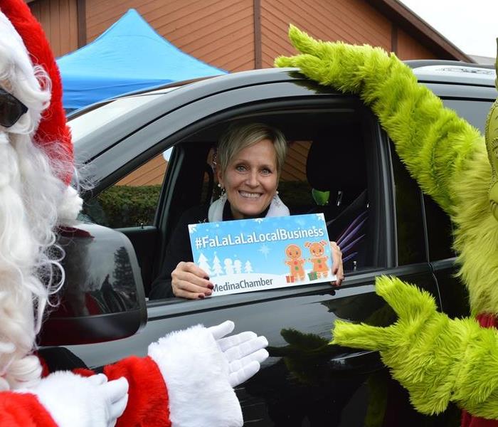 Female in a car holding sign for Medina Chamber and Santa Claus and the Grinch are standing outside of the car